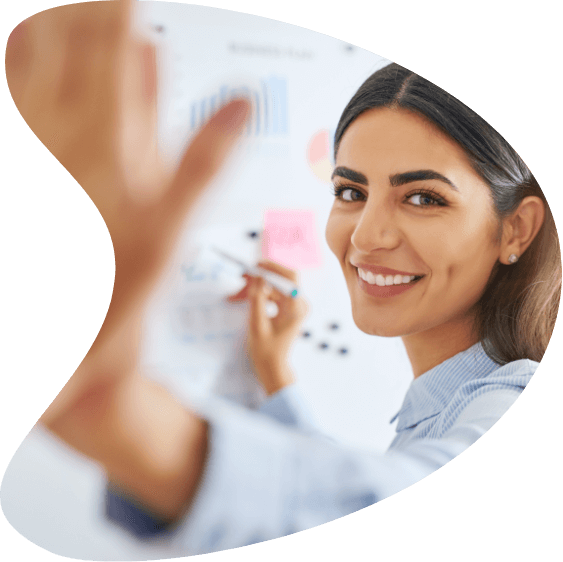 A woman smiling and standing in front of a whiteboard filled with charts and sticky notes, while writing with a marker, illustrates how organized planning can make jobs more efficient.