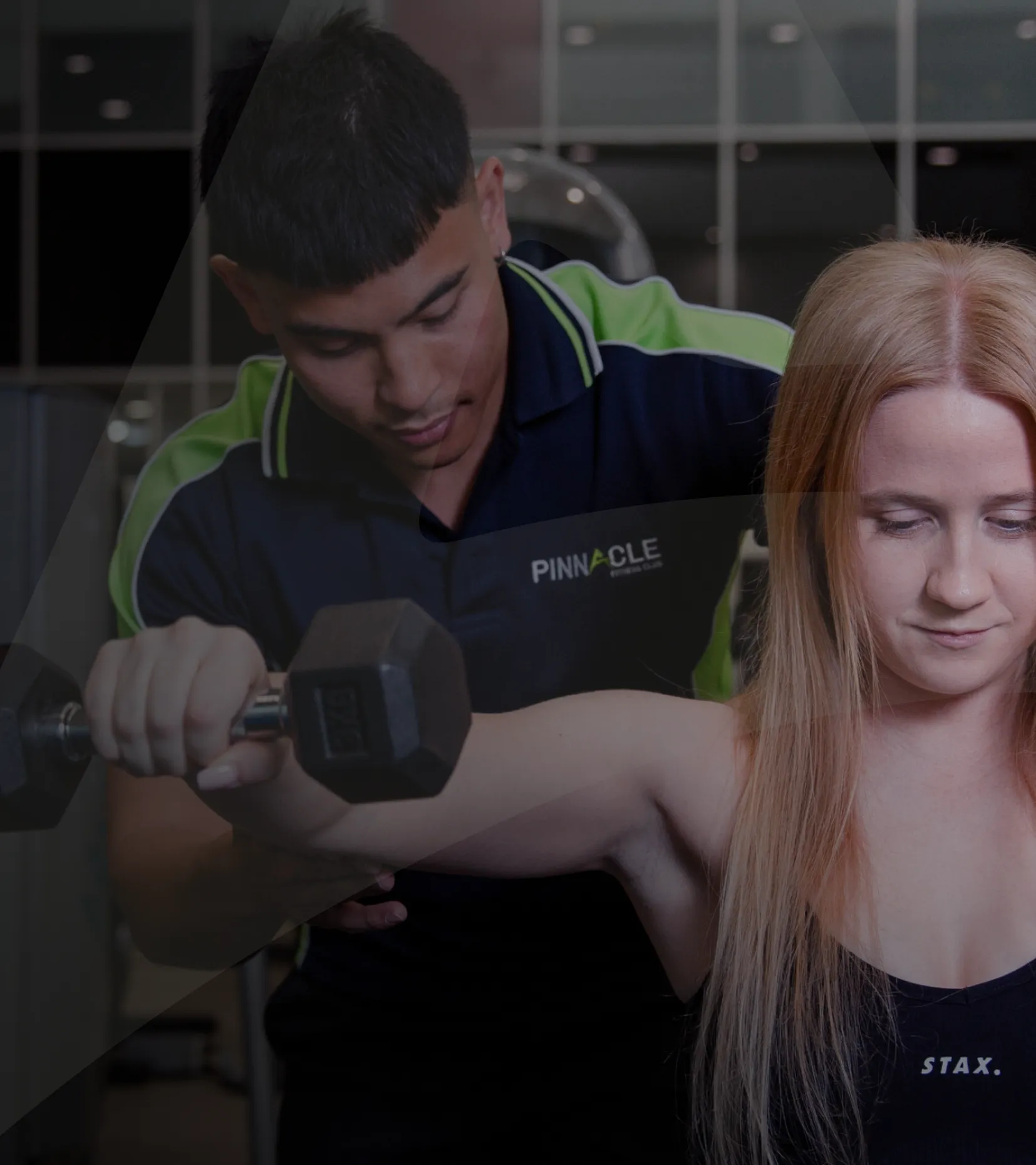 A personal trainer assists a woman lifting a dumbbell at a gym. The trainer wears a shirt with "Pinnacle" written on it, and the woman focuses on her strength training exercise.
