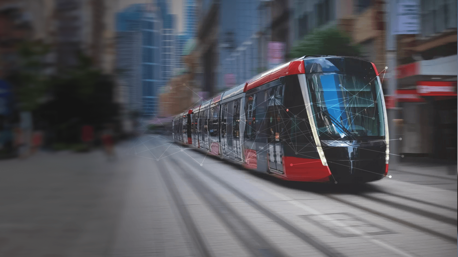 A modern, red and black tram moves along tracks through an urban city street flanked by tall buildings.