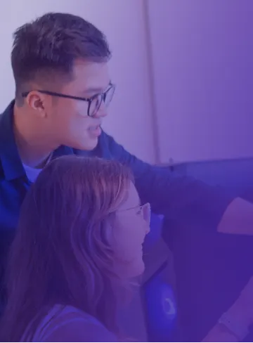 Two people wearing glasses are looking at a computer screen, with one pointing at the screen. The image is tinged with a purple hue.