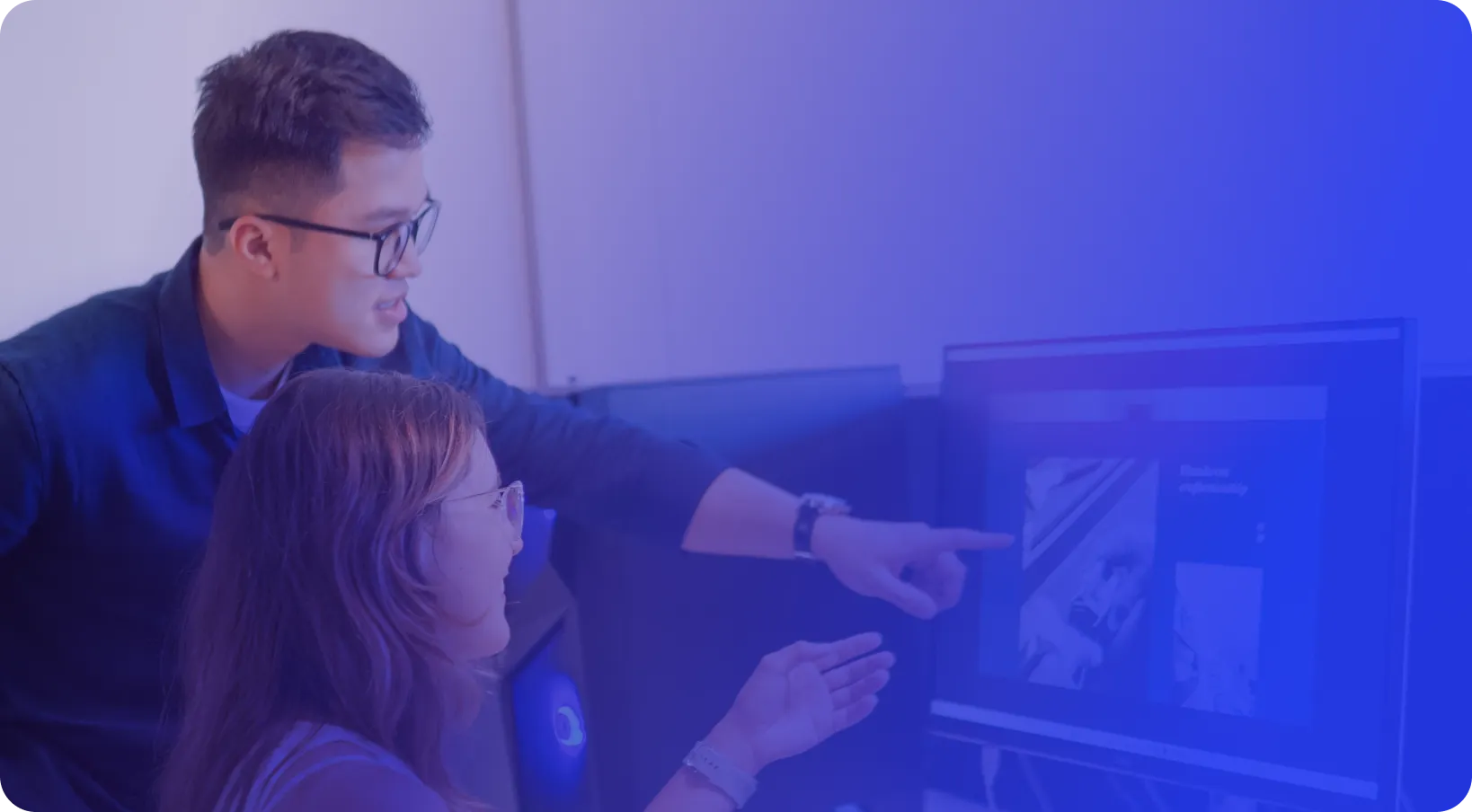 Two people, one standing and one sitting, look at a computer screen and discuss the content displayed. The scene is lit with a blue tint.